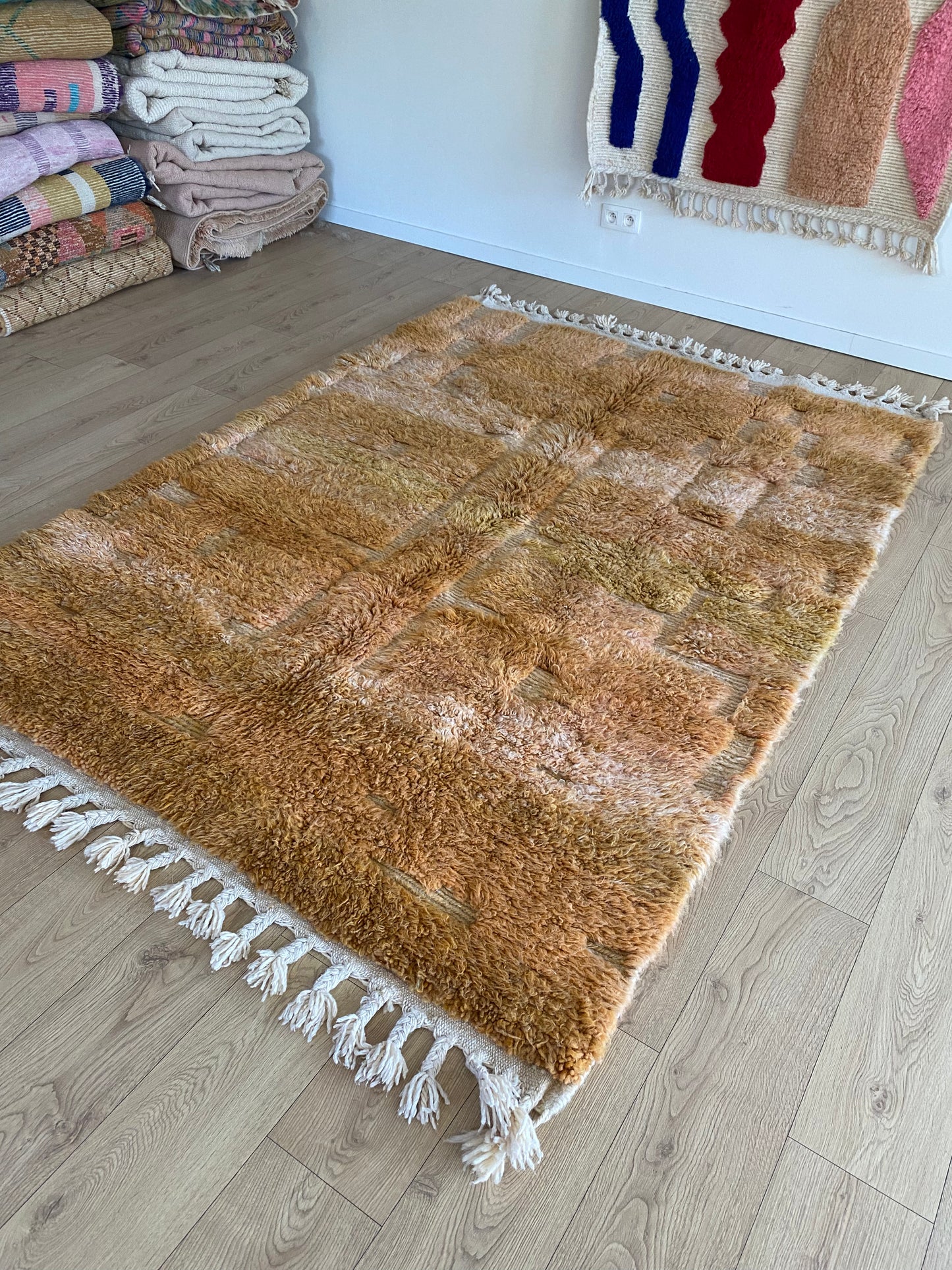 Brown textured rug with white tassels on a wooden floor, with stacks of folded fabric in the background.